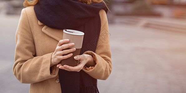 一个女人的手拿着一个穿着米色外套和黑色围巾的咖啡杯站在户外的画像 时尚的年轻女子穿着城市背景的秋装 着色图像图片
