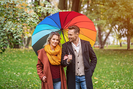 一对漂亮的年轻夫妇打着彩虹色的雨伞在公园里散步 一个漂亮的女孩在雨天和一个男人手挽着手穿过秋天的公园友谊季节家庭拥抱成人女朋友女图片