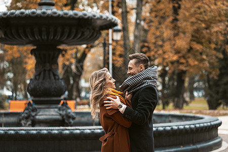 一对漂亮的年轻人站在秋天公园一个美丽的喷泉旁 穿着秋天的外套 互相拥抱 看着对方 着色图像公园城市夫妻女士女孩闲暇男朋友女朋友男图片