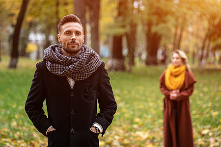 男人穿着外套和围巾站在公园里 等待站在他身后或从后面看起来像他的女朋友或妻子 美丽的秋日 年轻夫妇在公园里户外恋人享受女士成人夫图片