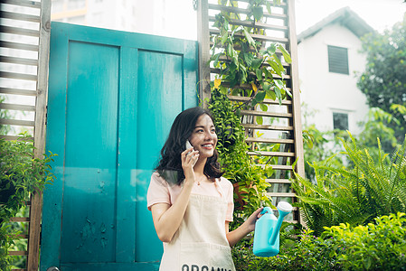 夏天在花园里浇花的亚洲女园艺人女性阳台手机微笑种植主妇植物花朵女孩房子背景图片