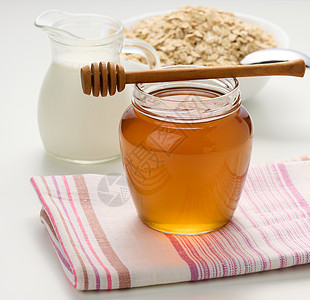 蜂蜜在玻璃透明罐子和白桌上的木棍上 在装有牛奶和燕麦的脱盐机后面图片