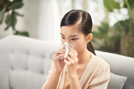 生病的年轻女人坐在沙发上 在家里的客厅里擤鼻涕 在纸巾打喷嚏的女人的照片流感过敏鼻音发烧药品感染疾病女士鼻子手帕图片