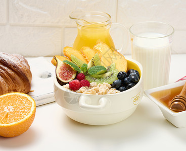 盘子里放着燕麦片和水果 半个成熟的橙子和鲜榨果汁放在透明玻璃瓶中 蜂蜜放在白桌上的碗里 健康早餐牛奶面包食物勺子羊角谷物陶瓷稀饭图片