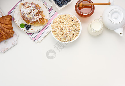 早餐早饭 陶瓷板上生燕麦片 脱甘油牛奶 蓝莓和白桌上的罐子蜂蜜餐巾面包饮料羊角甜点小吃毛巾杯子桌子食物图片