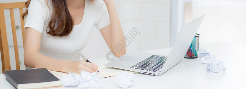 使用笔记本电脑工作的年轻亚洲女性认为创意项目和纸在家里的桌子上皱巴巴的有问题 女孩使用笔记本带着沮丧和麻烦 商业和自由职业者的概图片