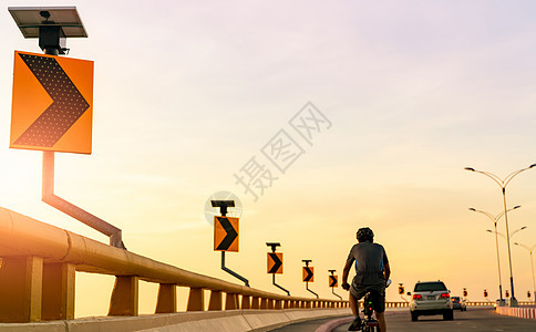 男人在汽车后面的弯道上戴着头盔骑自行车的背影 在路上安全驾驶和骑行 曲线与太阳能电池板能量的道路交通标志 户外运动 健康的生活方图片