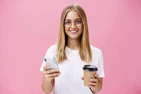 愉快友好的女孩微笑着拿着纸杯咖啡和智能手机的相机 快乐的好女人喝晨间饮料的肖像 在粉红色的墙上对互联网上的咖啡馆发表意见情绪广告图片