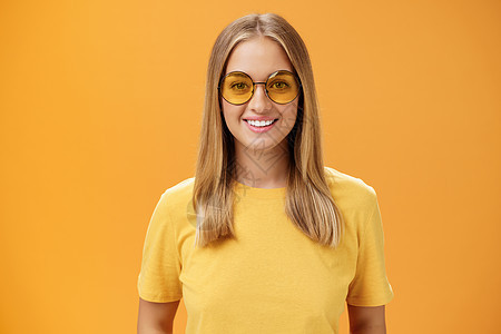 时尚自信迷人的欧洲女性 头发金黄 皮肤晒黑 身穿黄色 T 恤 戴着太阳镜 面带微笑 愉快地凝视着在橙色背景下摆姿势的相机框架青年图片