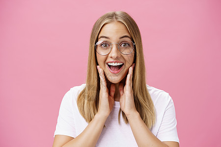 喜欢在店里挑选的新眼镜的女人感到很高兴找到合适的镜框抚摸着脸颊 幸福地对着镜头微笑 戴着透明眼镜和粉红色墙壁上的 T 恤金发女郎图片