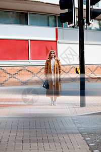 一名身穿棕色战壕的妇女站在一个行人交叉口等待城市交通灯光图片