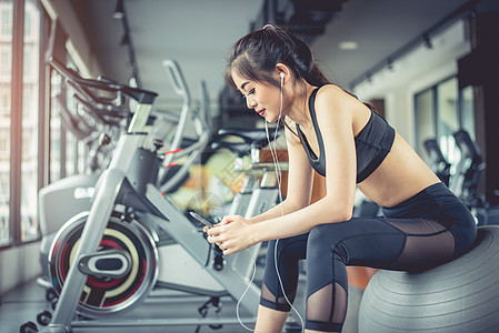 亚洲女性坐在健身球上 用智能手机在健身训练馆听音乐 放松和运动锻炼训练概念 科技娱乐主题图片