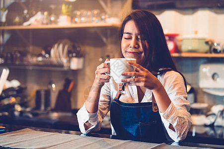 美丽的亚洲女咖啡师拿着一杯咖啡 品尝咖啡厅背景的煮咖啡 女服务员在自助餐厅闻咖啡 食物和饮料的概念 人们的生活方式图片