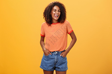 欢乐的热情和有魅力的年轻非裔美裔女性 有着ffro发型 快乐地挥舞着舌头 用手握在橘子墙口袋上 笑声爆炸牛角思维广告女朋友潮人橙图片