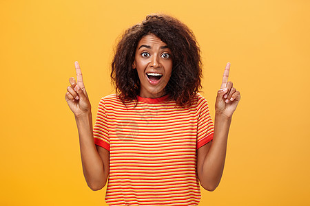 惊讶快乐热情可爱的非洲裔美国女性 头发卷曲 穿着条纹 T 恤 举起食指向上凝视 在橙色墙壁上看着相机 着迷和高兴潮人广告手势女朋图片
