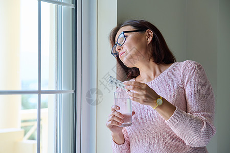 在家中手握着避孕药和水杯的中年妇女疾病玻璃女性药片药剂药物治疗镇痛药窗户成人图片