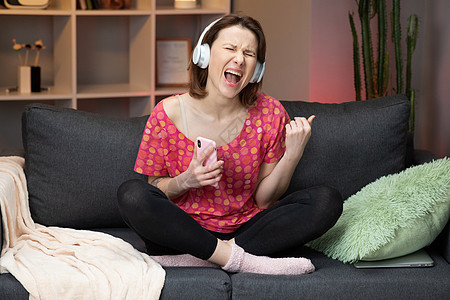 白人女孩坐在沙发上 笑着用现代智能手机在耳机中听音乐和微笑图片