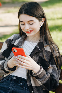 年轻美丽的女孩坐在公园的长椅上 在她的智能手机上通过社交网络磁带聆听音乐和叶子 (笑声)图片