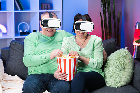 老白人配偶坐在沙发上吃爆米花 戴着 VR 护目镜看电影 家庭夫妇坐在沙发上吃爆米花 用 VR 眼镜看电视图片