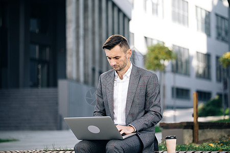 城里的年轻商务人士坐在长凳上 拿着笔记本电脑工作 在室外的便携式计算机上打字的商务人士 穿着西装坐在长凳上用笔记本电脑工作的男人图片