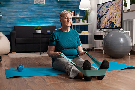 退休的老年妇女坐在瑜伽垫子上 用伸展弹性带伸展腿肌肉力量卫生健身房地面运动员运动身体客厅运动装锻炼图片