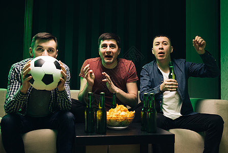 三个英俊快乐的白人友好男人坐在沙发上吃着薯条小吃 为电视上的足球比赛欢呼 看体育频道的男性朋友聚在一起图片