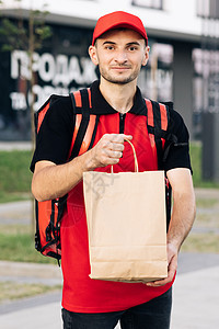 穿着红色制服的送货员拿着食品袋等待顾客的室外肖像 快乐的年轻快递员为他站在街上微笑的工作感到自豪 送货上门图片