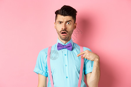 令人困惑的有趣的家伙在领结和吊带上 指自己好像被控告或被选中一样 引起眉毛惊讶 站在粉红背景旁站立商业胡子成人快乐办公室促销男人图片