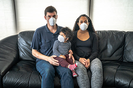 一个年轻的混血家庭坐在黑色真皮沙发上 抱着他们的儿子 脸上戴着防尘口罩 希望能防止因冠状病毒或 COVID-19 而生病或生病图片