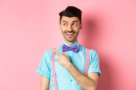 洋洋得意的男人 穿着有趣的领领 向左看 用左手瞄准 展示宣传标志 站在粉红背景上吊带裤标识广告办公室领结商业胡子成功促销微笑背景图片