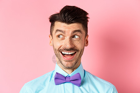 特写笑得开朗的男人 看着一脸快乐的标志 查看很酷的促销优惠 站在粉红色背景下成人工作室商业办公室成功吊带裤领结发型胡须胡子图片