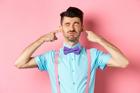 戴着领结的恼怒的滑稽男人闭上耳朵 塞住手指 做鬼脸 被巨响激怒 站在粉红色的背景上胡须发型工作室成功办公室成人情绪商业促销快乐图片