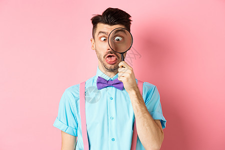 穿领领领带的有趣男人 通过放大玻璃看 斜视和做傻脸 站在粉红背景上工作室微笑成功发型放大镜吊带裤快乐办公室促销成人图片