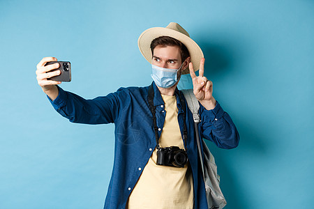 Covid19 大流行病和旅行概念 在旅游帽中快乐而积极的家伙拿自拍和展示和平标志 摆近观光的蓝色背景社交帽子假期乐趣生活保健广图片