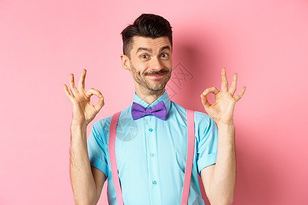 留着胡子和领结的帅哥的画像 摆出不错的手势 微笑着表示赞同 推荐好的报价 站在粉红色的背景上商业男人情绪快乐成功吊带裤发型工作室背景图片
