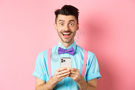 阅读智能手机屏幕 在相机上微笑 站在粉红背景面前的欢笑和兴奋的人 他们眼中的喜悦之人男人胡须技术成功广告工作室工具办公室成人快乐图片