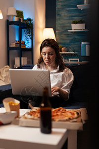 妇女一边看纪录片 一边在笔记本电脑上工作 一边吃着美味的零食小吃垃圾食物啤酒瓶子记录沙发喜剧面条午餐图片