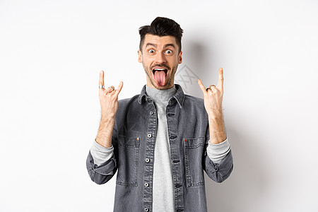令人兴奋的幽默的家伙 展示舌头和重金属角标志 享受派对或活动 玩乐 站在白色背景上男人发型广告胡子快乐胡须商业情绪工作室微笑图片