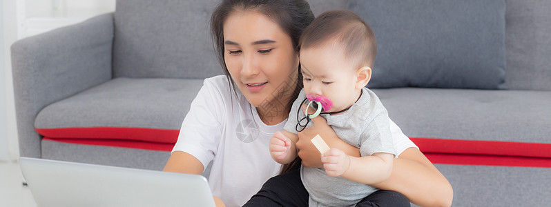 年轻的亚洲母亲在笔记本电脑上工作 同时在家照顾女儿 妈妈和女婴坐在一起使用笔记本视频通话 父母和亲密关系 室内 家庭和商业概念幸图片