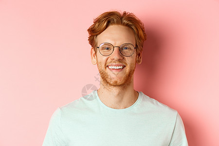 特写快乐的红头发男人的脸 对着镜头微笑着用白牙 戴着眼镜以获得更好的视力和 T 恤 站在粉红色的背景上成人发型学生胡子购物工作室图片