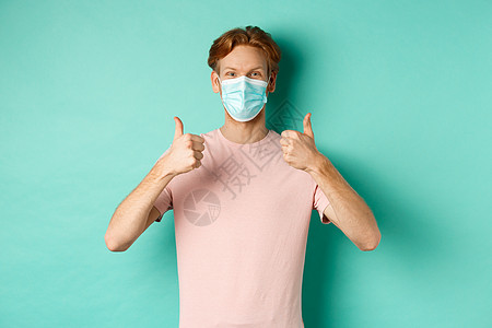Covid19 大流行病和生活方式概念 在医疗面具中装着快乐的红发人 站在绿松石背景上站立 举起大拇指来表示赞美及赞美产品商业医图片