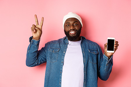 在线购物和技术概念 快乐的黑人在蜜蜂和牛仔衬衫上 展示移动屏幕与和平标志 站在粉红色背景面部站立情感工作室互联网手机冒充潮人电话图片