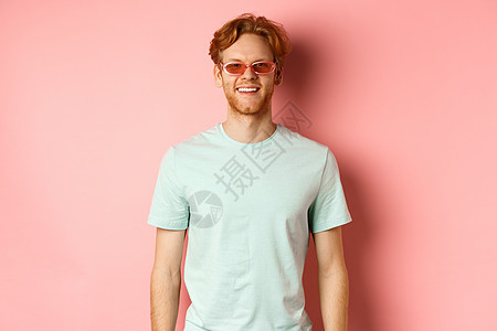 旅游和度假的概念 戴着墨镜和 T 恤 欢快的红发大胡子男子 站在粉红色背景上 对着镜头微笑 看起来很开心头发成人冒充太阳镜眼镜广图片