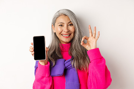 时尚的亚洲高龄女性显示空白智能手机屏幕和OK标志 微笑喜悦 推荐应用程序 白背景等   info whatsthis奶奶促销购物图片