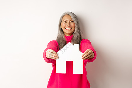 快乐的韩国中年妇女 带着灰色头发 展示纸房模特和微笑 穿着粉红色毛衣站在白底背景上老年女性广告购物派对房地产化妆品房子奶奶公寓背景图片