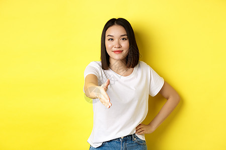 自信的亚洲女性穿着白色 T 恤 伸出手握手和打招呼 打招呼 自我介绍 站在黄色背景上手势快乐情感广告工作室成人黑发青少年发型女孩图片