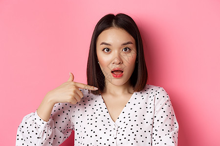 困惑的亚洲女性站在粉红色背景上 指着自己 垂下下巴 盯着镜头提问的形象黑发女孩微笑冒充购物质疑促销广告学生情感图片