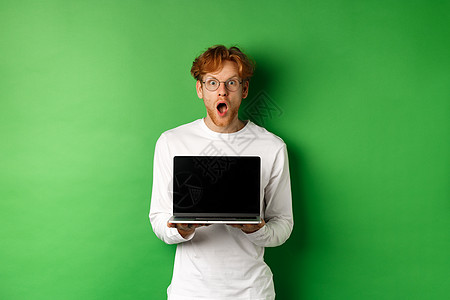 戴着眼镜的红头发男子惊讶地看到笔记本电脑空白屏幕 显示在线宣传和下巴 盯着相机惊叹 绿色背景胡须男性学生购物自由职业者广告商业展图片