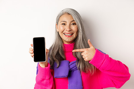 美丽的亚洲祖母微笑 指着智能手机空白屏幕 显示移动应用程序 站在白色背景上    action inlife beautiful图片