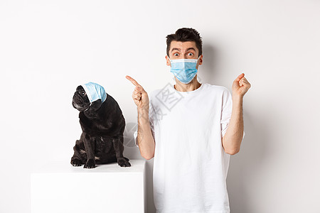 Covid19 动物和检疫概念 戴着医用口罩的年轻人和黑狗 哈巴狗看着左上角 老板展示促销优惠 欣喜若狂快乐潮人小狗男性工作室社图片